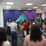 A group of people stand in an office room, engaged in a discussion, with desks and computers in the background.