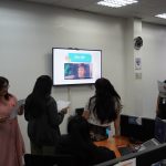 A group of people in an office setting watch a presentation on a wall-mounted screen. Some are holding papers. The room has a few desks and a clock on the wall.