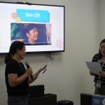 Two people stand in front of a TV screen labeled "Video Clip," holding microphones and papers. They are smiling and appear to be engaged in a presentation.