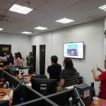 Office meeting with several people standing and looking at a presentation on a screen. Desks with computers are in the foreground.