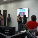 A group of people stand in an office room, some holding papers. A screen on the wall displays a presentation.