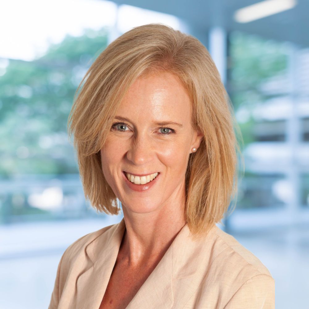 Delia MacKenzie, a woman with shoulder-length blonde hair, wearing a beige blazer, smiles in an indoor setting with a blurred background.