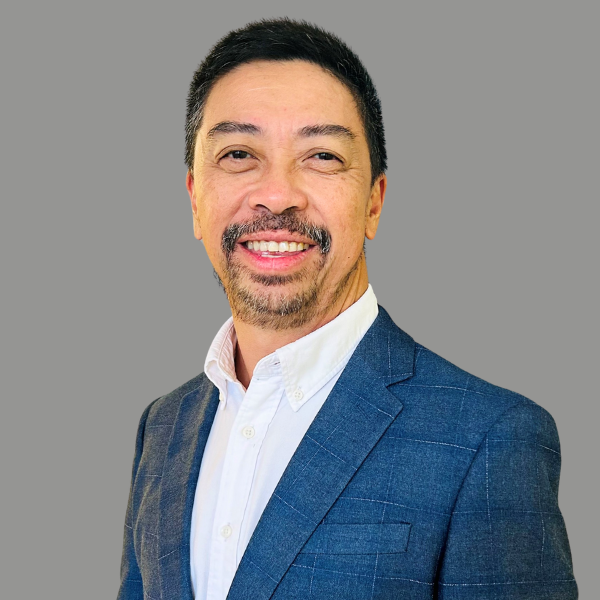 A man with short dark hair and a beard is wearing a blue suit jacket and white shirt, smiling against a plain gray background, ready to announce the PM 10th Anniversary Conference in 2024.