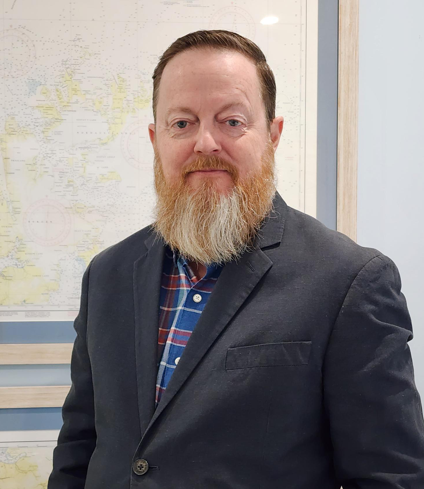 A man with a beard wearing a plaid shirt and blazer poses for a portrait in front of a framed map on the wall, commemorating the 10th Anniversary of Profitmaster at the conference.